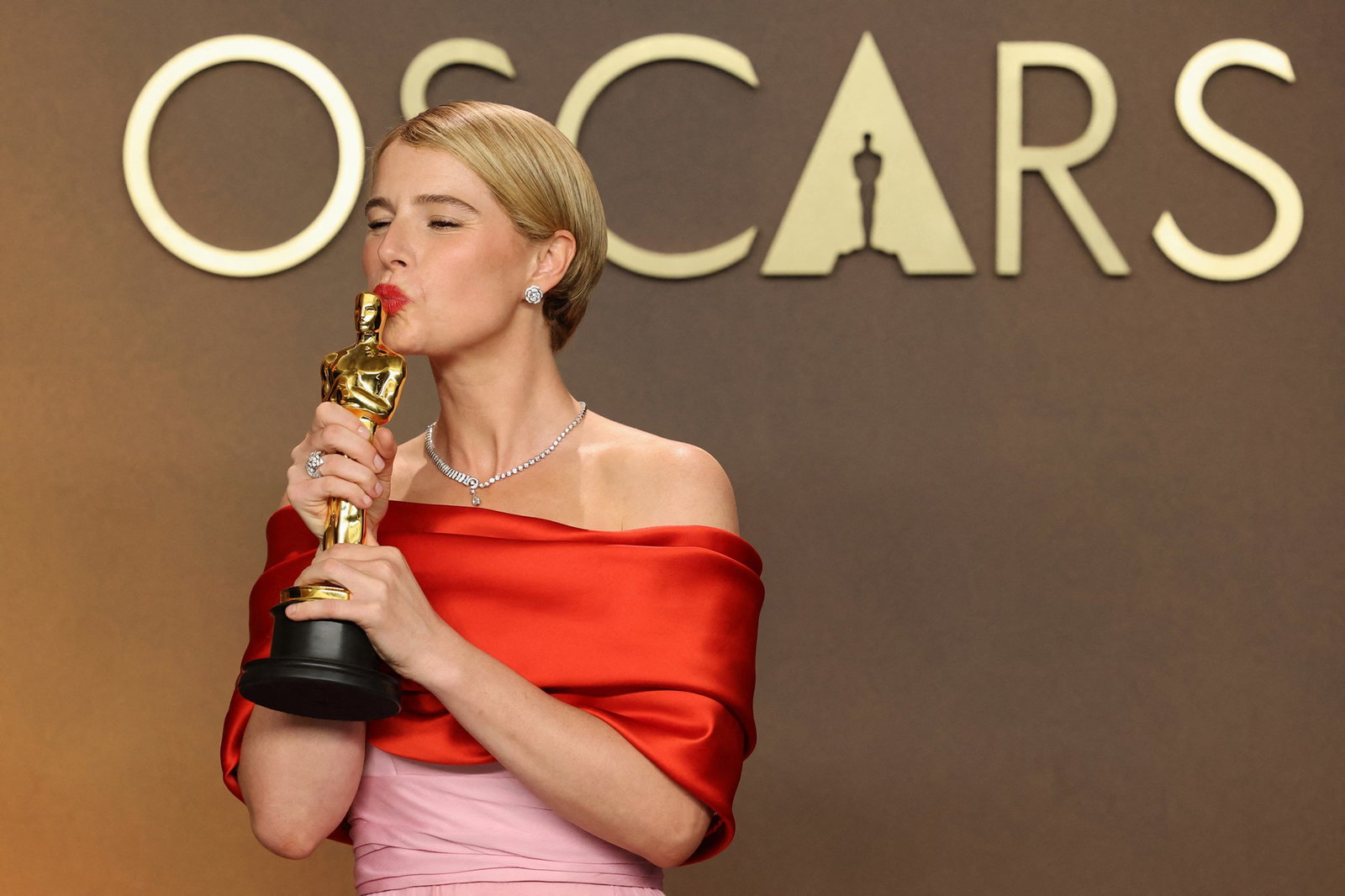 A woman in a red and pink dress kisses the top of her gold Oscar.