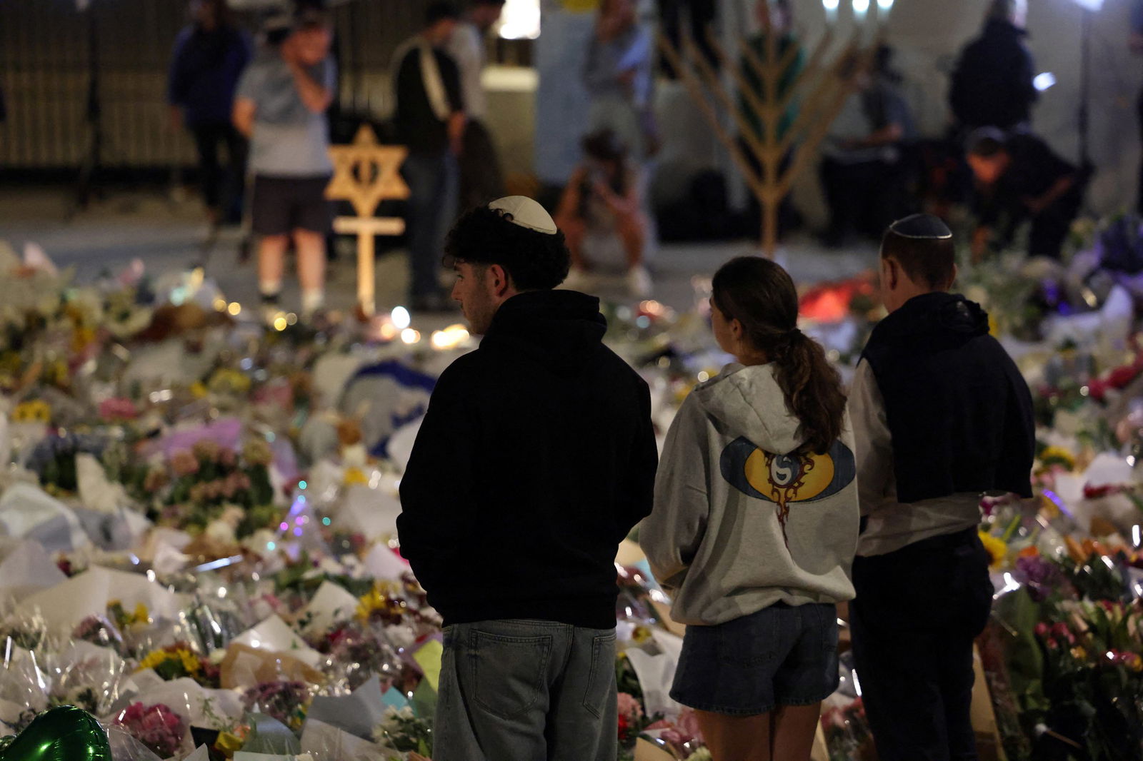 Três pessoas olham para uma grande pilha de flores e homenagens.