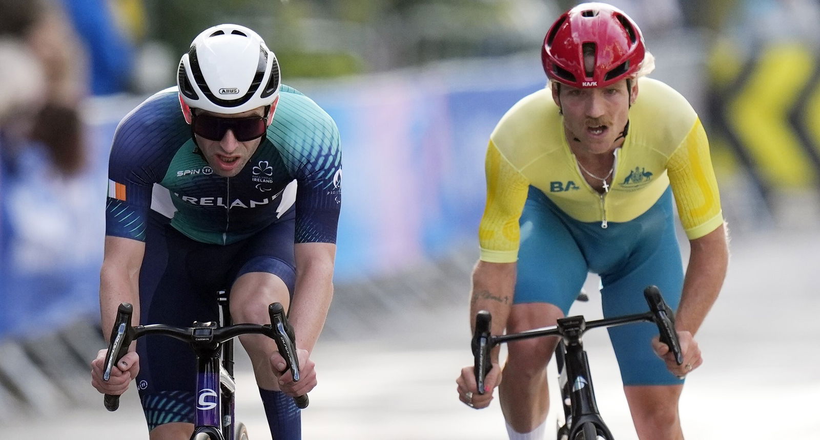 A cyclist in green and gold is next to a cyclist in blue and green. They are riding bikes and looking forward with their mouths open