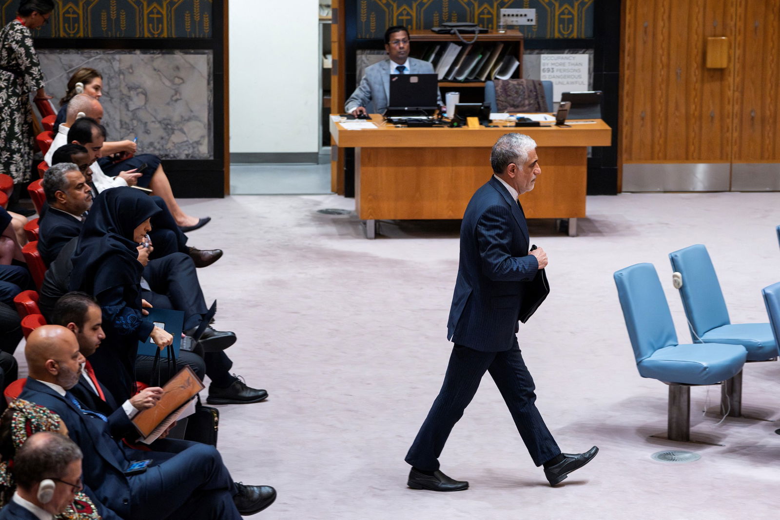 A man in a suit walks from a seated group of people to a set of blue chairs
