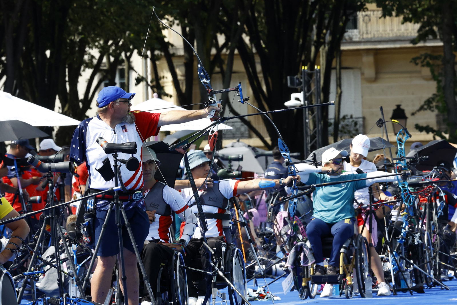 Male para-archers are lined up with one standing and three others in wheelchair, shooting during competition.