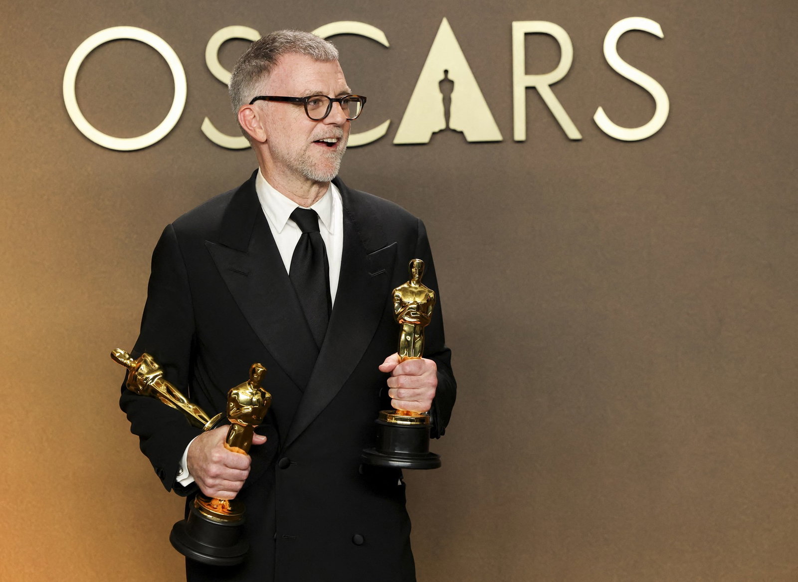 A man in a black suit and tie holds three gold Oscars, and smiles.